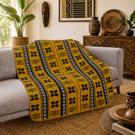 A mustard yellow throw blanket with black and white African wax print patterns is draped over a light gray sofa in a styled living room setting.