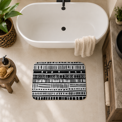 A rectangular bath mat with a black and white geometric tribal pattern is placed on a beige tiled floor in front of a white freestanding bathtub. A cream towel is draped over the tub, with a small wooden stool, plant, and bathroom decor visible around it.