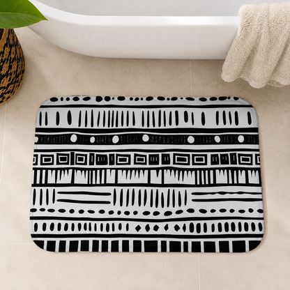 A rectangular bath mat with rounded corners featuring a bold black and white geometric tribal pattern, styled on a beige tiled bathroom floor next to a white bathtub with a folded beige towel.