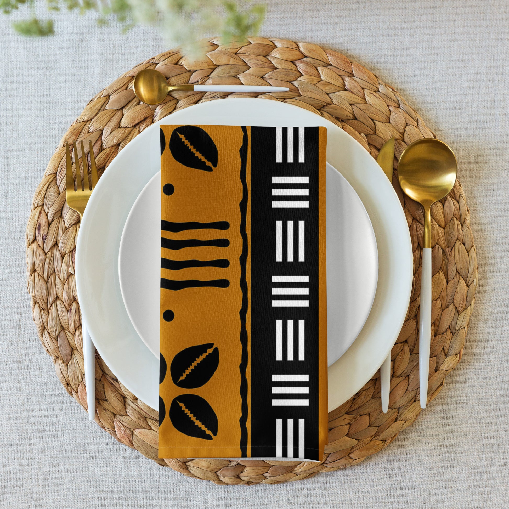 A napkin with a black and gold geometric pattern on a round white plate, with a gold fork and spoon on either side, placed on a woven placemat.