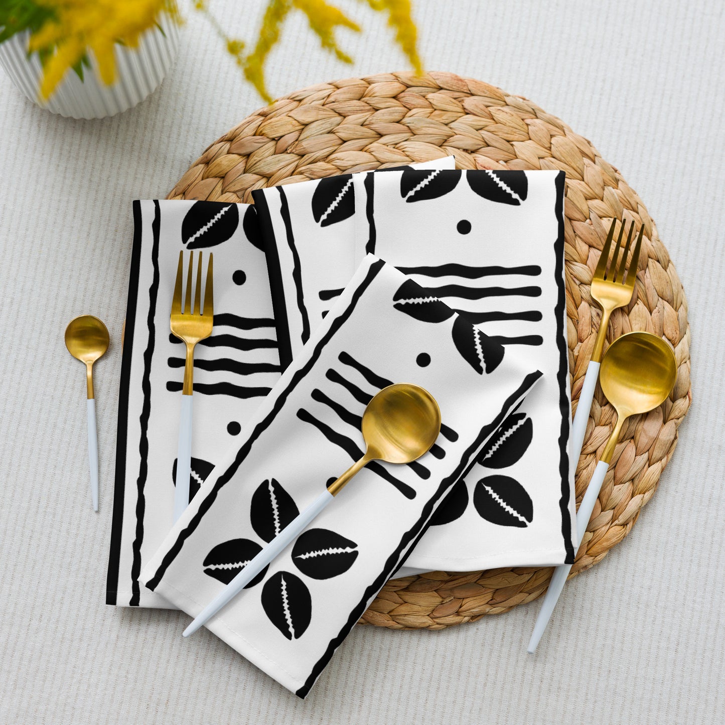 A set of white cloth napkins with a black African shell pattern, displayed on a woven placemat with gold-colored cutlery.