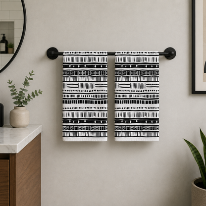 Two black and white patterned towels hanging from a black metal towel bar on a beige bathroom wall, with a vanity, mirror, and plants visible in the room.