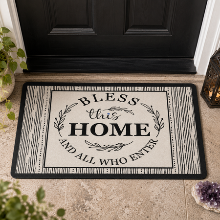 Doormat with 'Bless this Home and All Who Enter' text on a black and white design, placed in front of a black door.