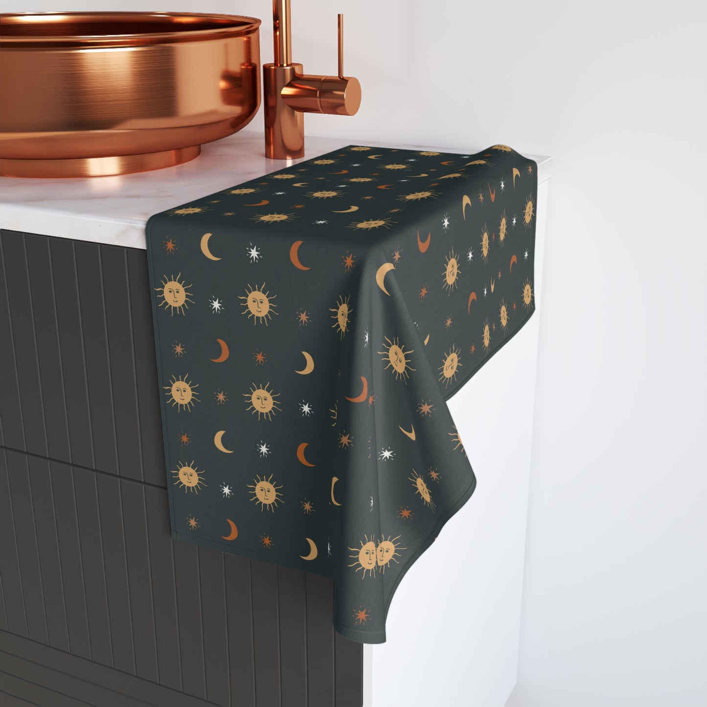 Dark green decorative hand towel with celestial pattern draped over a table next to a copper sink.
