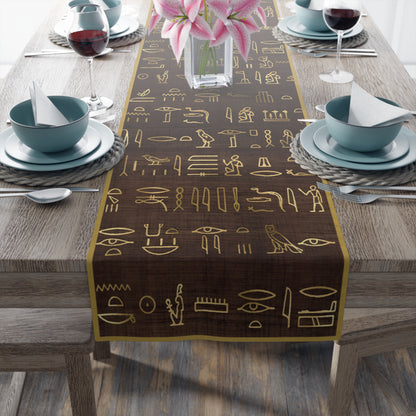 A table runner with Egyptian hieroglyphics printed in brown color, laid out on a wooden dining table with plates and glasses set on top.