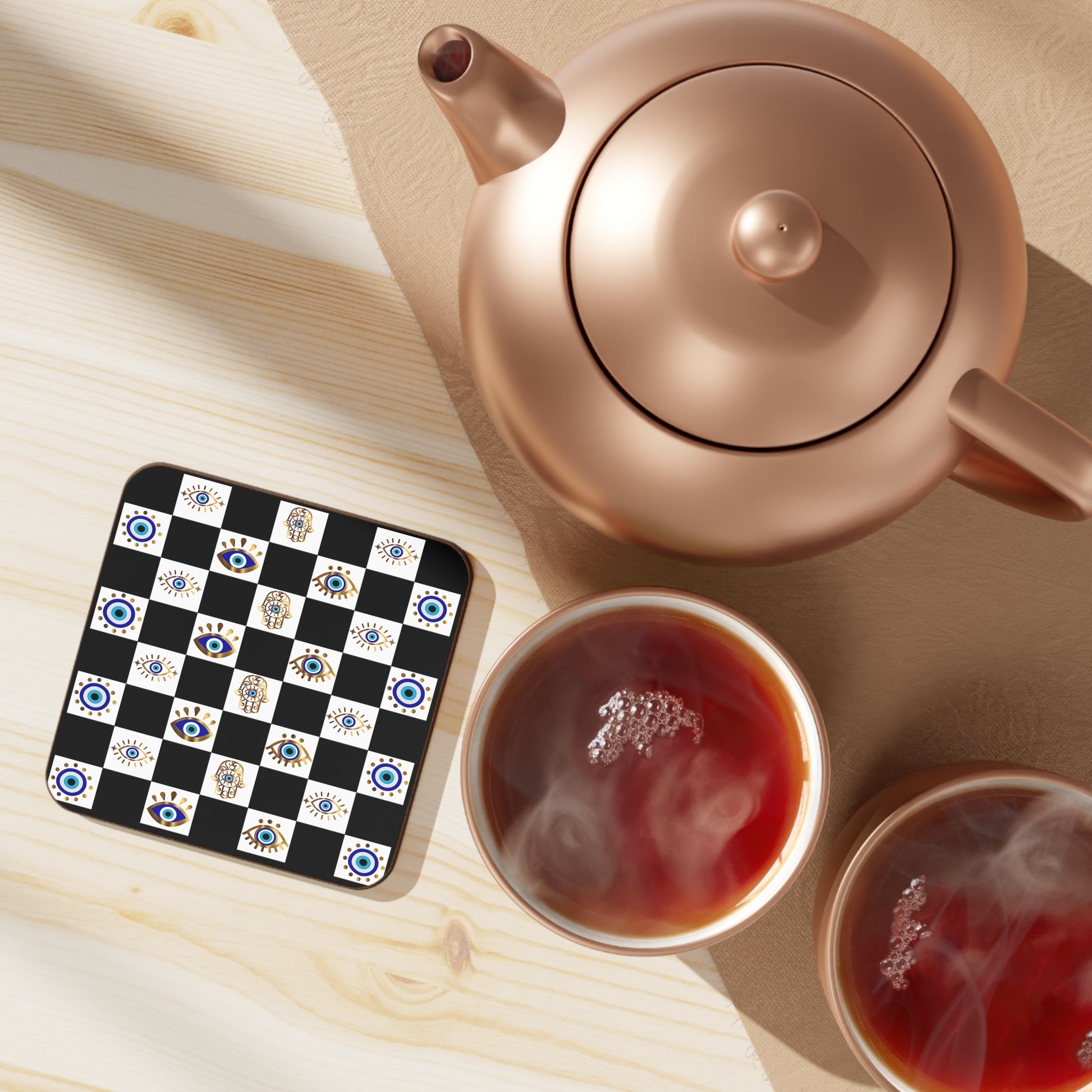 Rose gold teapot with two cups of tea on a checkered evil eye hamsa coaster set.