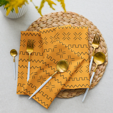 A yellow napkin with a geometric mudcloth design placed on a white plate, with a gold fork and spoon on either side, set on a woven placemat.