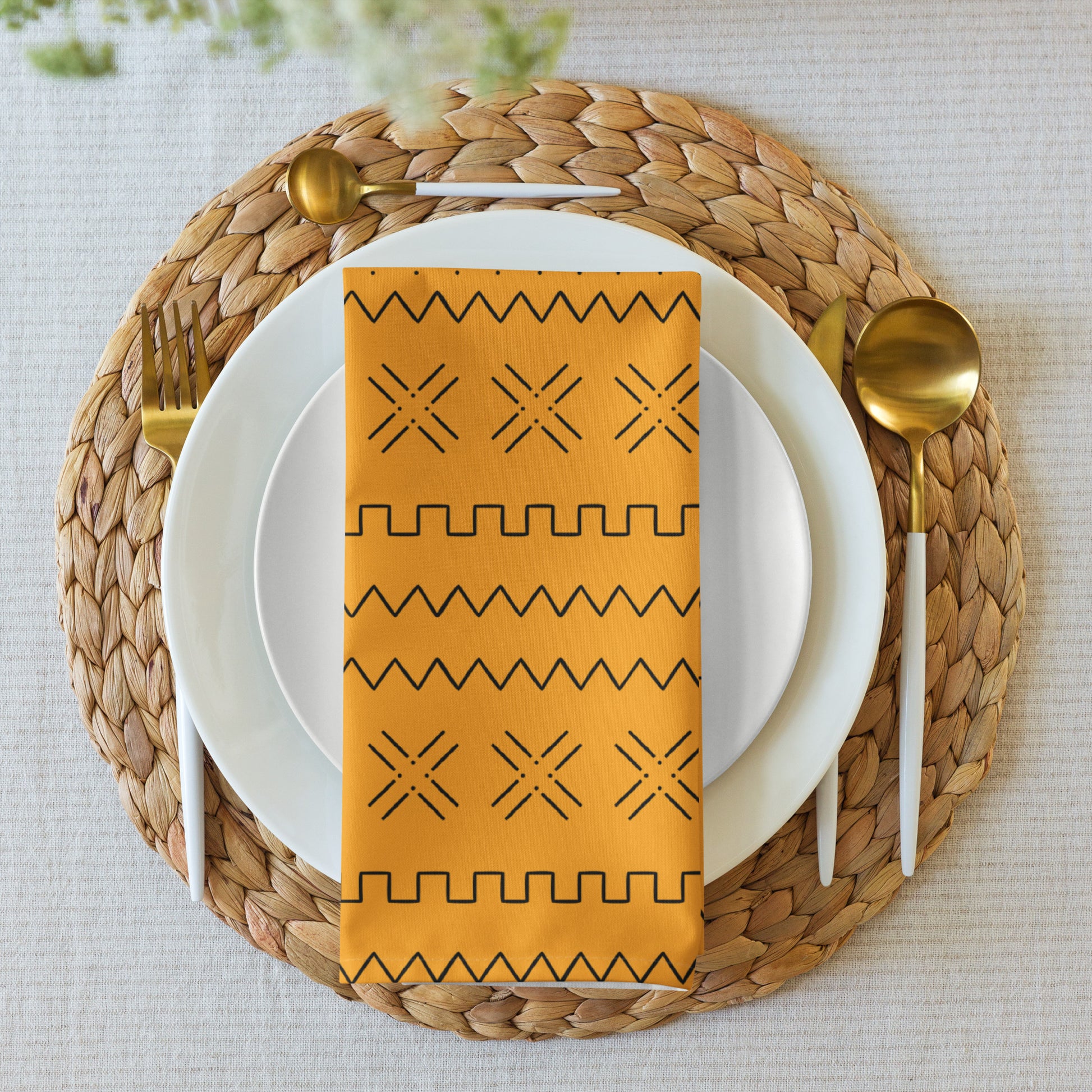 A yellow napkin with a geometric mudcloth design placed on a white plate, with a gold fork and spoon on either side, set on a woven placemat.