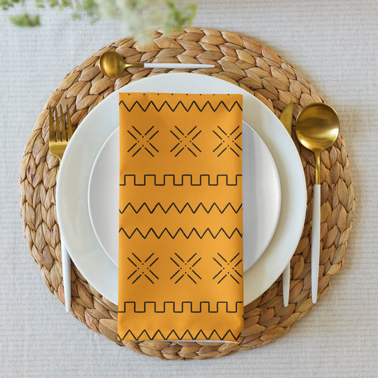 A yellow napkin with a geometric mudcloth design placed on a white plate, with a gold fork and spoon on either side, set on a woven placemat.