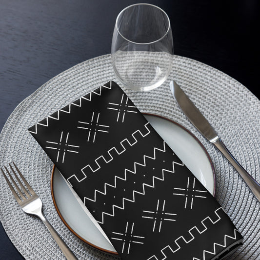 A black and white geometric mudcloth patterned napkin is displayed on a white plate with a glass and cutlery, placed on a woven grey placemat.