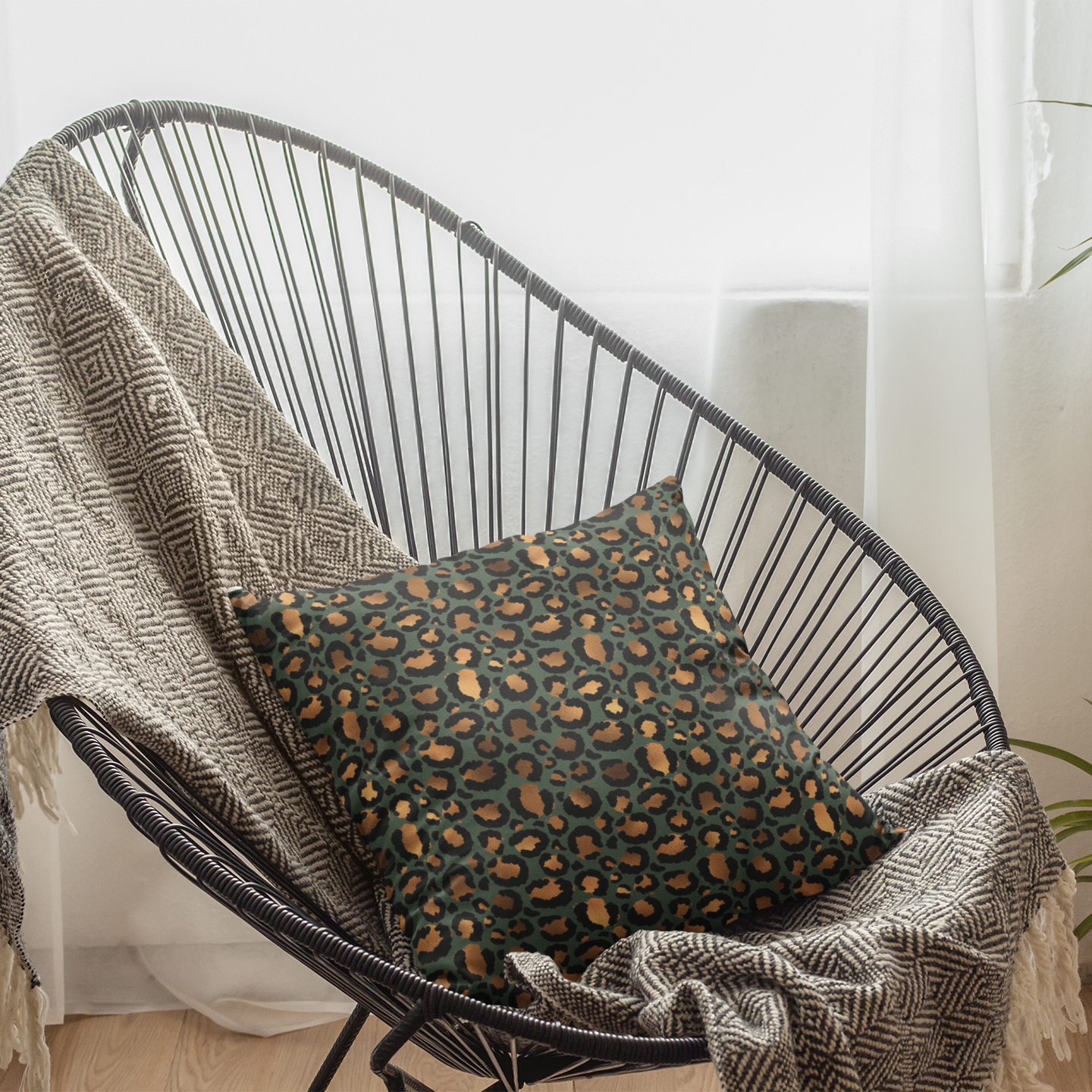 A leopard print throw pillow with a geometric pattern, draped over a black wire chair with a plant and a white curtain in the background.