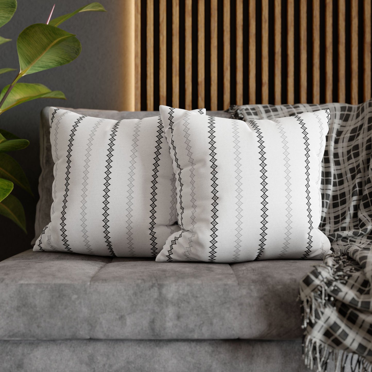 Two square throw pillows with a white mudcloth print on a gray couch, with a wooden slat background and a green plant to the side.