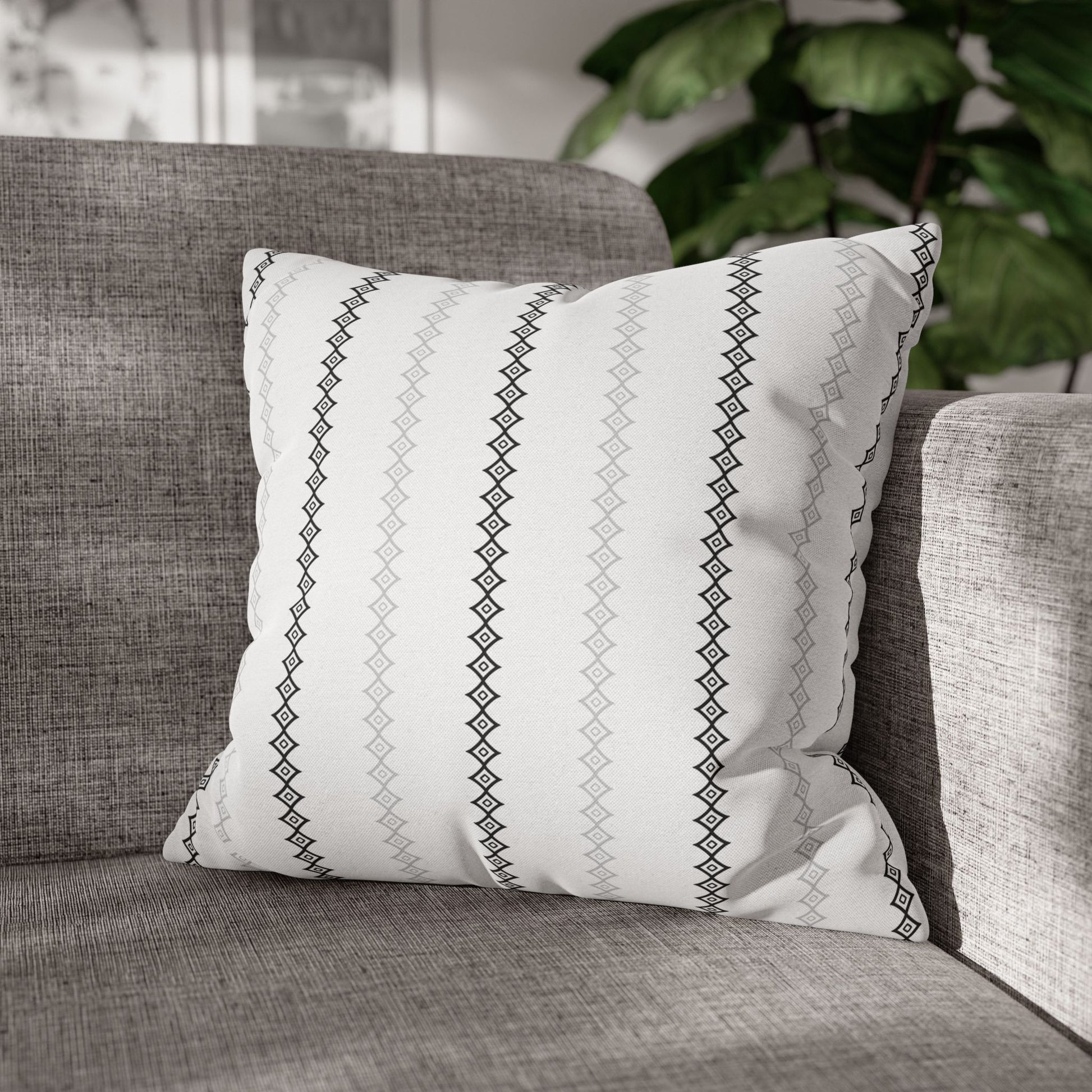 Two square throw pillows with a white mudcloth print on a gray couch, with a wooden slat background and a green plant to the side.