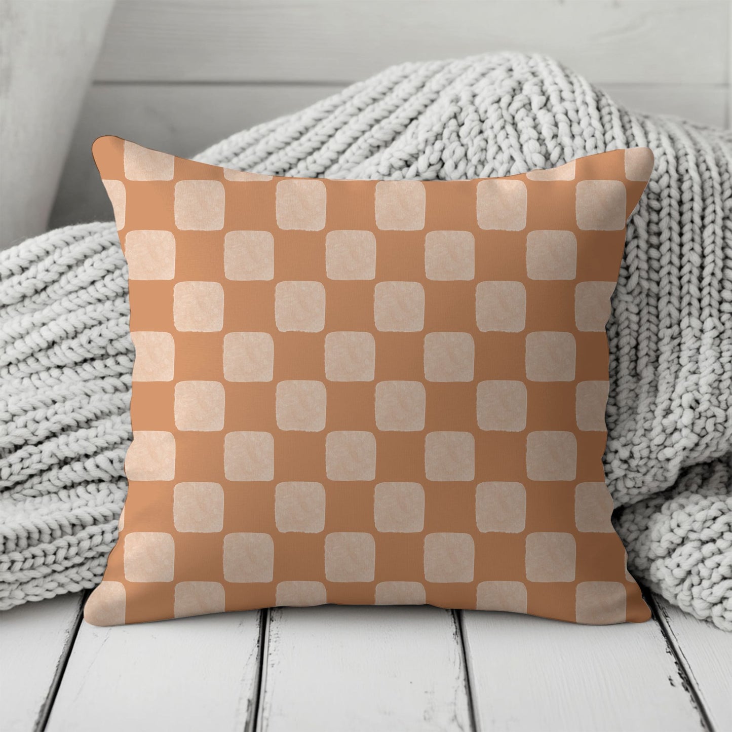 Checkered pillow with a peach brown and beige pattern on a wooden surface with a textured gray blanket.
