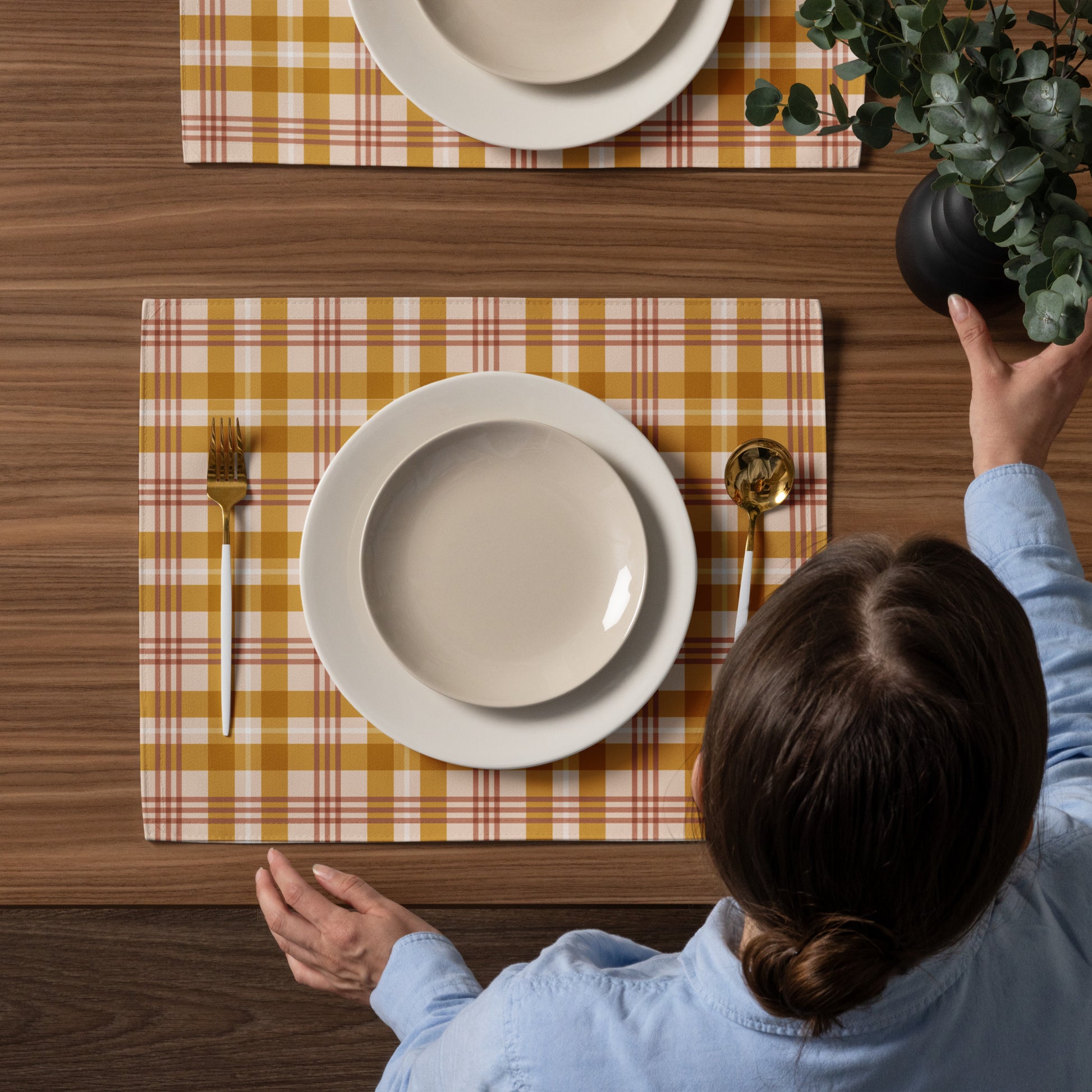 Person setting a table with yellow plaid placemats and white plates.