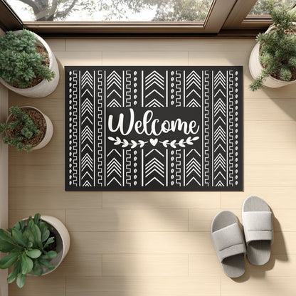 Decorative doormat with 'Welcome' text and Mudcloth patterns on a wooden floor with plants and slippers.