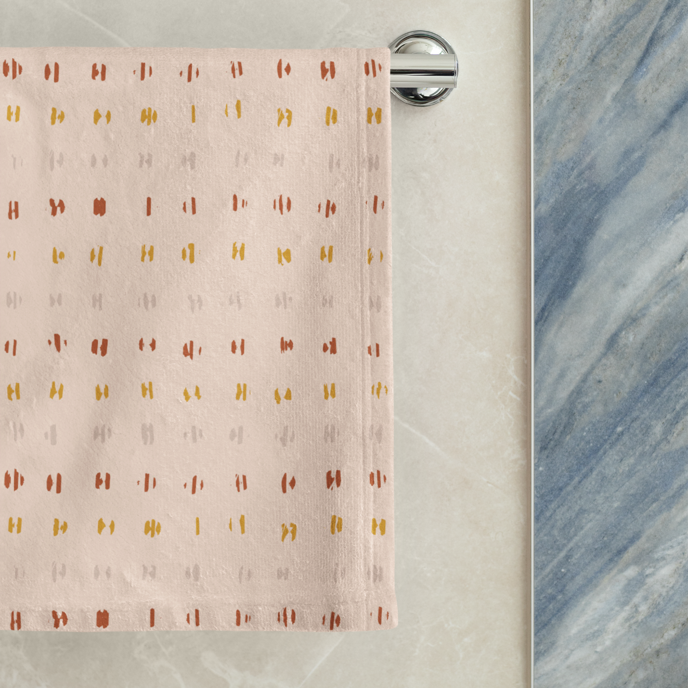 Pink towel with gold geometric pattern hanging on a silver towel rack against a marble wall.