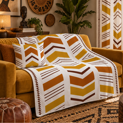 A cozy living room scene featuring a mustard yellow armchair draped with a white throw blanket and matching pillow, both printed with a bold chevron pattern in mustard yellow, terracotta orange, and brown. The room is styled with warm bohemian decor, including a woven pendant light, a leather pouf, and patterned wall art.